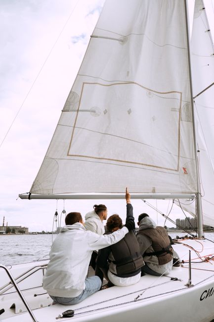 Image of a white sailing boat with three crew members on board, navigating through choppy waters near a large bridge. One crew member is sitting outside the boat, holding onto the side, while another 
